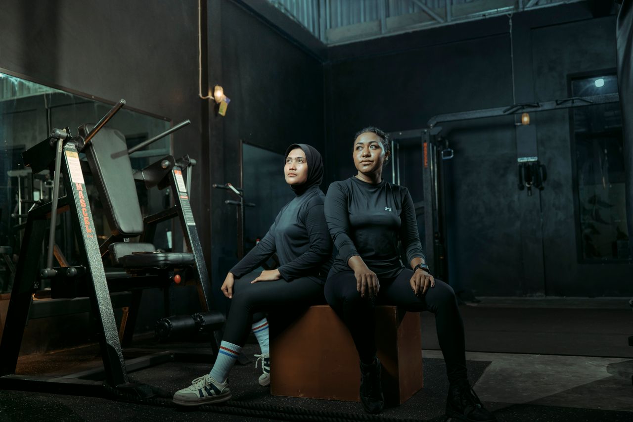 Classes Two women in athletic wear sitting on a box with gym equipment in the background.